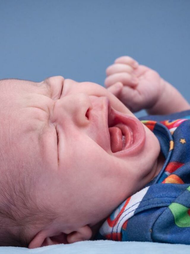 Bebê deitado com uma roupa azul com detalhes coloridos, chorando bastante