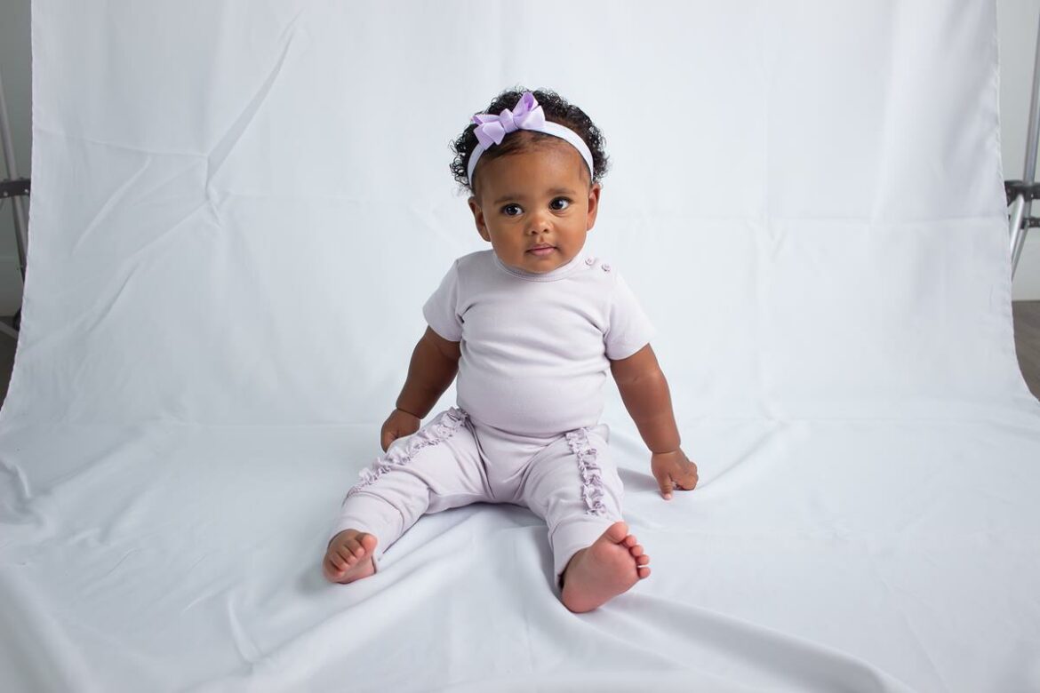 Bebê sentado sobre fundo branco, usando conjunto lilás com detalhes de babados e faixa de cabelo lilás com laço, olhando atentamente para frente.