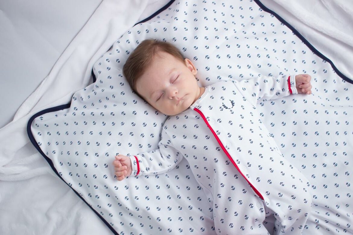 Bebê dormindo tranquilamente de macacão branco com estampa de âncoras azuis e detalhes vermelhos, deitado sobre manta combinando.