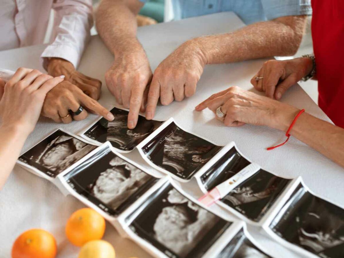Várias mãos apontam com carinho para imagens de ultrassonografias dispostas sobre a mesa, representando um momento de união familiar e emoção diante da chegada de um novo bebê.