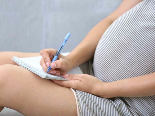 Mulher grávida sentada fazendo anotações em um caderno com uma caneta azul, possivelmente organizando uma lista de presentes para o chá revelação.