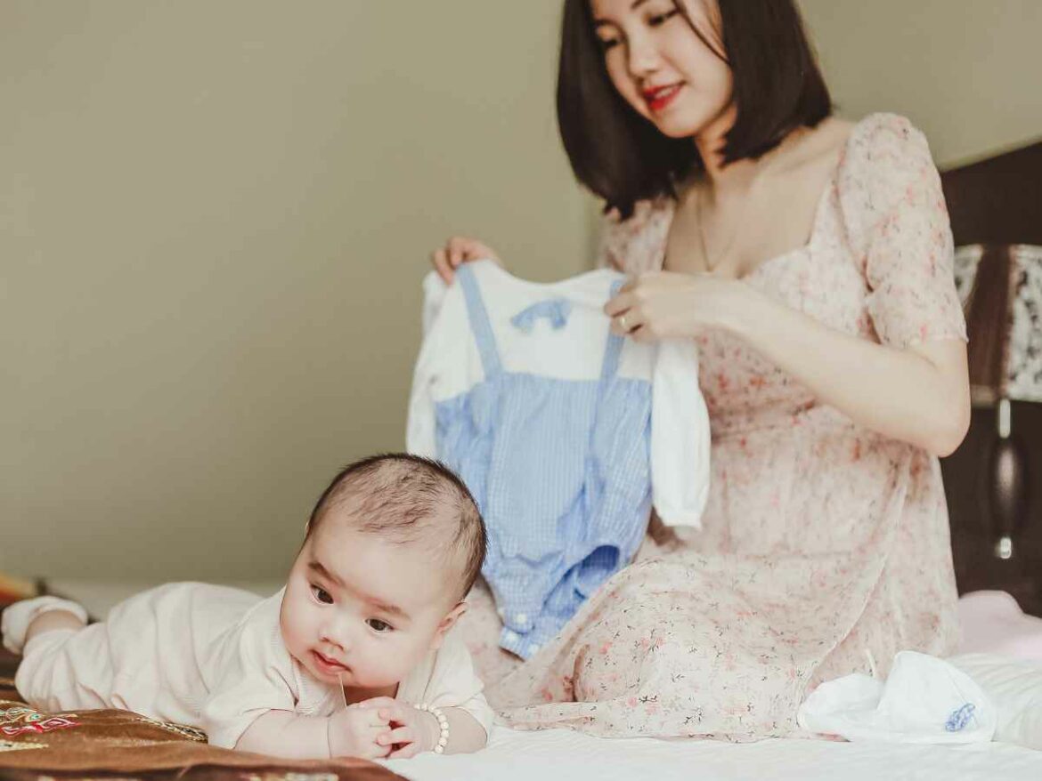 Mãe sentada em uma cama segura um macacão azul e branco enquanto observa seu bebê deitado de bruços ao lado dela, pronto para a troca de roupa.