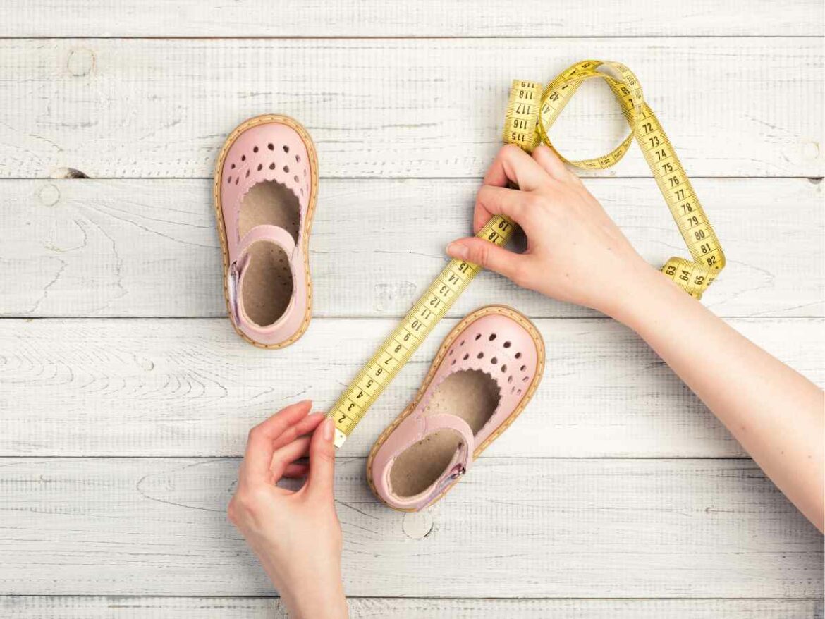 Mãos adultas medem com fita métrica o comprimento de sapatinhos infantis rosa sobre superfície de madeira clara