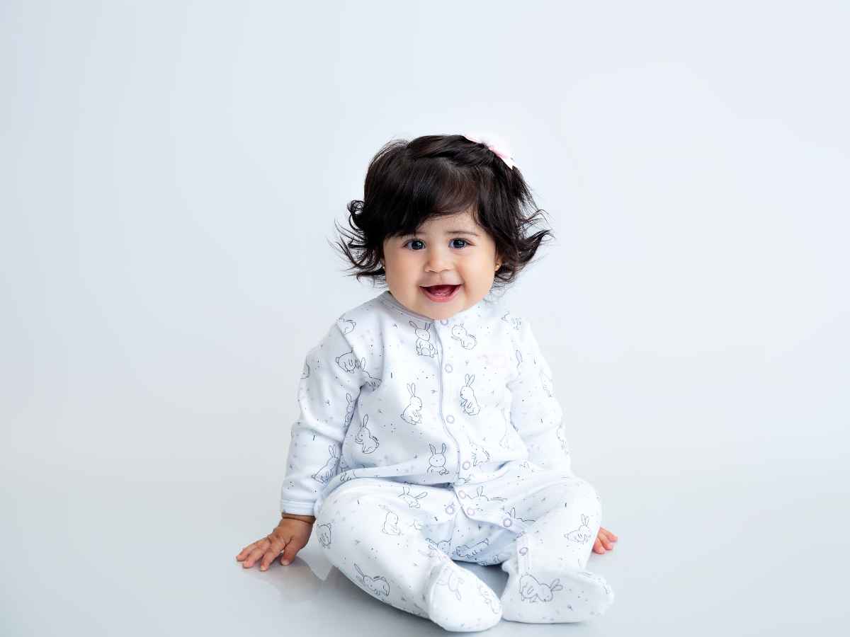 Bebe trajada com roupinhas de páscoa, ela usa um macacão branco com desenhos de coelhinho em azul claro. Fundo azul claro sólido.