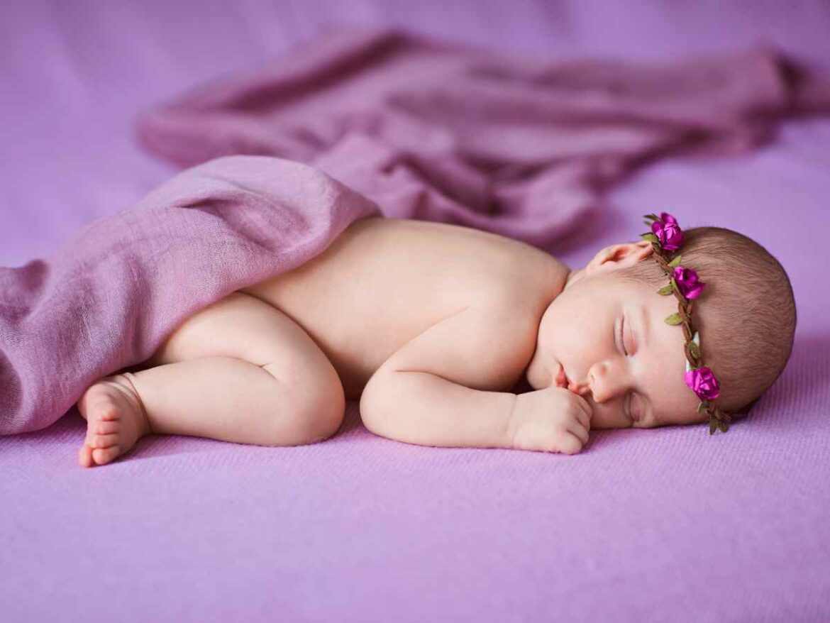 Bebê recém-nascido dormindo em posição fetal sobre manta lilás, usando faixa de flores roxas na cabeça e coberto parcialmente por tecido da mesma cor.