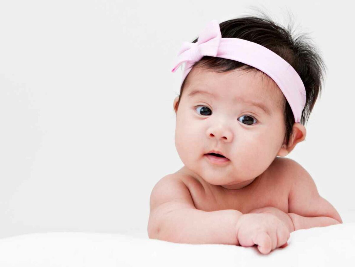 Bebê deitado de bruços com faixa rosa na cabeça, olhando curioso para a câmera em fundo branco.
