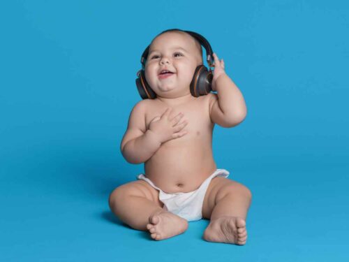 Bebê com expressão de felicidade usando fones de ouvido enquanto escuta música, transmitindo alegria e encantamento ao interagir com os sons. Fundo azul sólido.