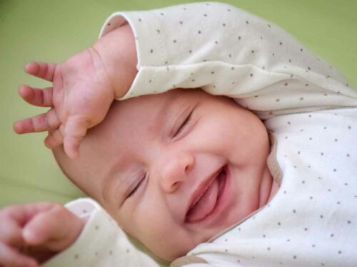 Bebê deitado, sorrindo e erguendo os braços com entusiasmo ao escutar música, reagindo de forma positiva e expressiva às sensações provocadas pelos sons, em um momento de alegria e estímulo emocional.