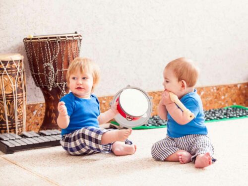 Dois irmãos bebês explorando sons enquanto brincam com instrumentos musicais. Um segura um pandeiro e o outro uma flauta, demonstrando curiosidade e diversão ao descobrir os diferentes sons que cada instrumento produz.