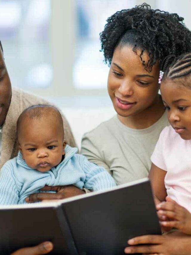 Casal de pais lendo a história do livro que escreveram especialmente para seus filhos pequenos. A mãe segura uma menina de cerca de 3 anos no colo enquanto segura o livro com a outra mão; ao lado, o pai segura um bebê de aproximadamente 8 meses e apoia o livro com a outra mão. Juntos, compartilham o momento de leitura em família, criando memórias afetivas.