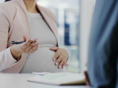Gestante vestindo um terno rosa, com a mão apoiada na barriga, gesticulando enquanto conversa com seu chefe sobre a importância de se desligar totalmente do trabalho para aproveitar plenamente o período da licença-maternidade.