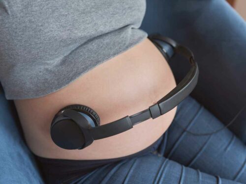 Mulher grávida posicionando fones de ouvido sobre a barriga para que seu bebê comece a se acostumar com sons e vibrações, promovendo estímulos sonoros ainda durante a gestação em um momento de conexão e cuidado.