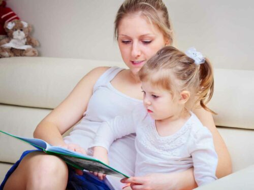 Mãe segurando sua filha no colo enquanto lê um livro que escreveu em homenagem a ela. A história tem poucos conflitos, mas é envolvente e encantadora, prendendo a atenção da menina, que escuta com interesse.
