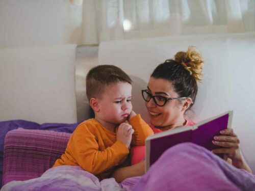 Mãe e filho aconchegados debaixo de uma coberta roxa, enquanto ela lê uma história em voz alta para criar um momento especial de conexão, carinho e afeto entre os dois.