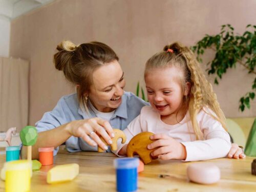 Mãe estimulando sua filha com atraso de desenvolvimento por meio de uma brincadeira com peças de madeira. As duas sorriem durante a atividade, criando um momento de aprendizado, conexão e alegria.
