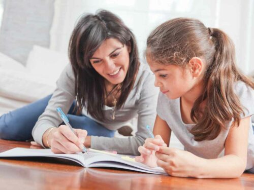 Mãe e filha deitadas no chão, sorrindo e envolvidas na criação de uma história especial para o bebê da família. Juntas, escrevem em um caderno com lápis, compartilhando imaginação, carinho e criatividade nesse momento em família.