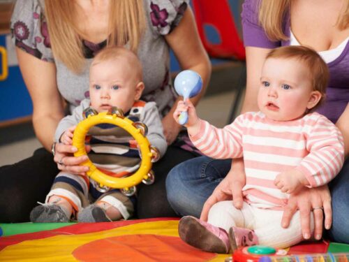Mães segurando seus filhos enquanto eles tocam instrumentos musicais juntos — um bebê com um chocalho e o outro com um pandeiro. A atividade promove o desenvolvimento cognitivo e emocional dos pequenos, fortalecendo o vínculo com suas mães por meio da música e da interação.