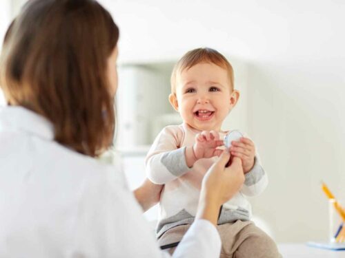 Médica de costas realizando uma avaliação clínica em um bebê para investigar possíveis causas de atraso de desenvolvimento. Ela segura um estetoscópio à frente do bebê, que sorri em resposta, criando um ambiente acolhedor e tranquilo durante o exame.