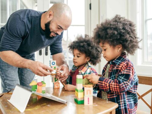Pai brincando com seus filhos gêmeos utilizando blocos de montar, com o objetivo de estimular a coordenação motora. Ele participa ativamente da atividade, promovendo desenvolvimento e vínculo afetivo de forma lúdica e educativa.