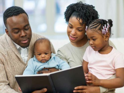 Casal de pais lendo a história do livro que escreveram especialmente para seus filhos pequenos. A mãe segura uma menina de cerca de 3 anos no colo enquanto segura o livro com a outra mão; ao lado, o pai segura um bebê de aproximadamente 8 meses e apoia o livro com a outra mão. Juntos, compartilham o momento de leitura em família, criando memórias afetivas.