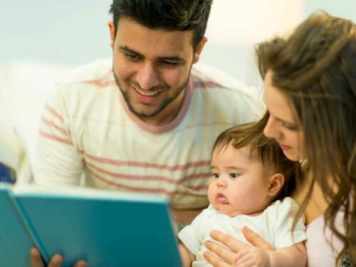 Pais lendo para seu bebê com o objetivo de fortalecer o vínculo emocional. A mãe segura a criança no colo com carinho, enquanto o pai segura um livro de capa azul e lê em voz alta, criando um momento de conexão e afeto em família.