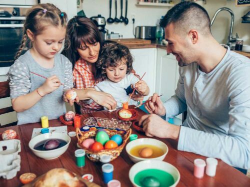 Família reunida celebrando a Páscoa com alegria e união. A mãe segura o bebê no colo enquanto o pai e a irmã ajudam a pintar ovos decorativos, todos envolvidos na atividade com carinho e entusiasmo.