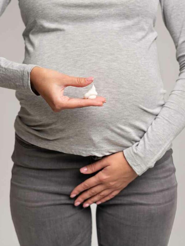 Grávida observando atentamente um corrimento em sua mão, preocupada com a ardência que tem sentido na região íntima, possivelmente relacionada a uma infecção urinária. Com a outra mão, ela toca a área da calça onde sente desconforto. Fundo cinza sólido.