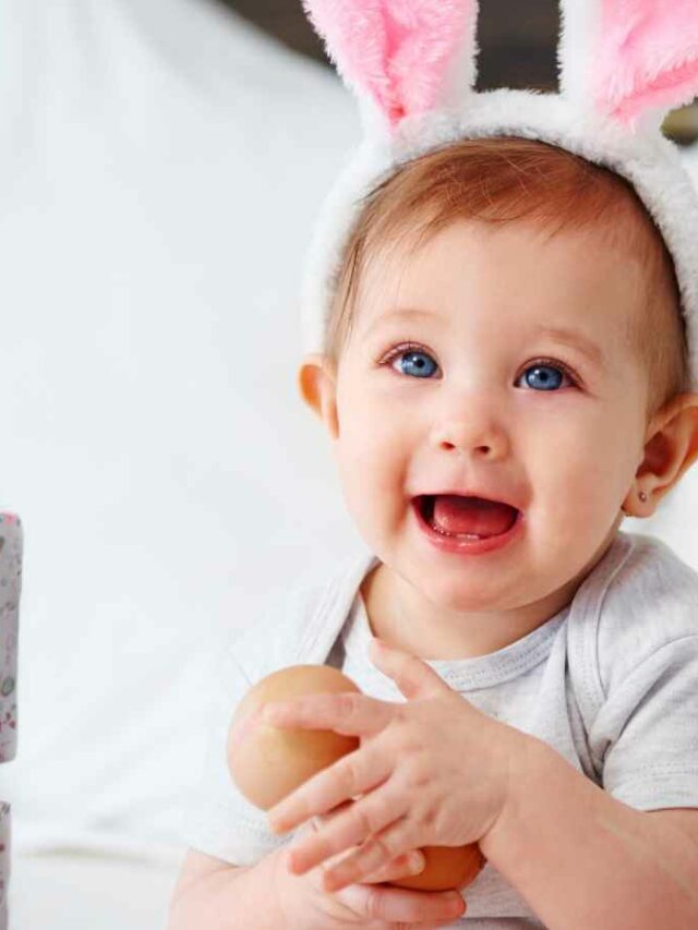 Bebê usando orelhas de coelho, sentado ao lado dos presentes que recebeu de Páscoa. Enquanto sorri.
