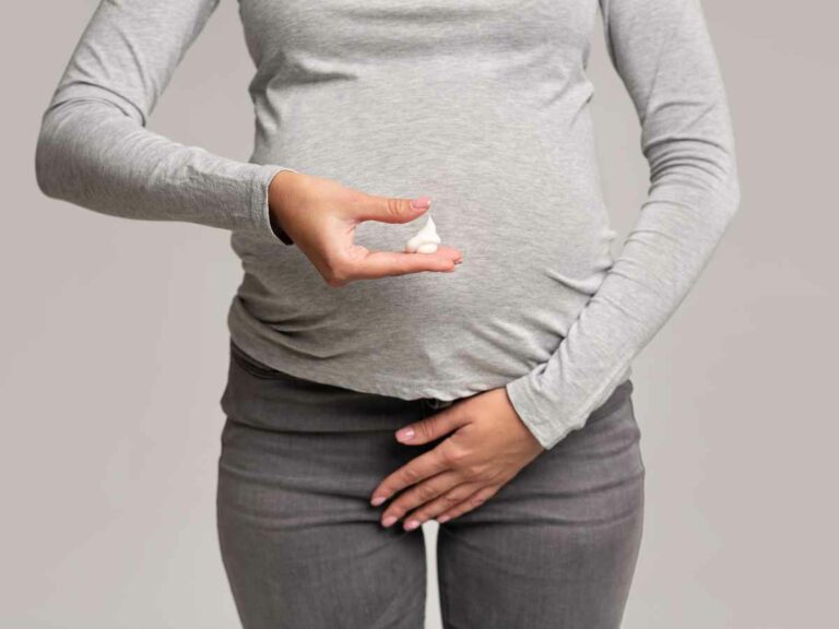 Grávida observando atentamente um corrimento em sua mão, preocupada com a ardência que tem sentido na região íntima, possivelmente relacionada a uma infecção urinária. Com a outra mão, ela toca a área da calça onde sente desconforto. Fundo cinza sólido.
