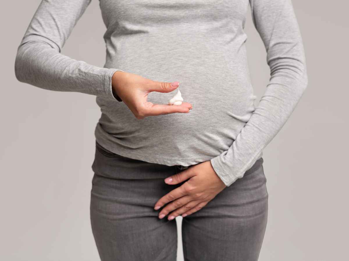 Grávida observando atentamente um corrimento em sua mão, preocupada com a ardência que tem sentido na região íntima, possivelmente relacionada a uma infecção urinária. Com a outra mão, ela toca a área da calça onde sente desconforto. Fundo cinza sólido.