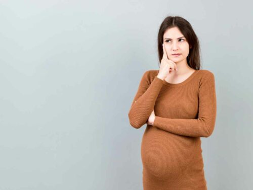 Grávida com expressão de dúvida no rosto, posando com a mão no queixo enquanto reflete sobre os sintomas que está sentindo. Ela está incerta se os sinais estão relacionados a uma infecção urinária ou vaginal. Fundo cinza sólido.