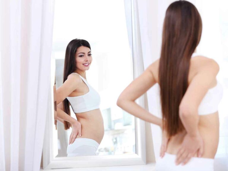 Mulher no puerpério se olhando no espelho, usando roupas íntimas brancas e observando atentamente seu corpo após a gestação. A imagem simboliza o momento de reflexão e aceitação das mudanças corporais que ocorrem no pós-parto.