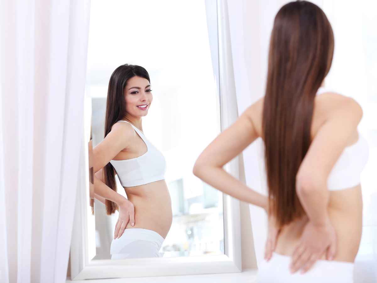 Mulher no puerpério se olhando no espelho, usando roupas íntimas brancas e observando atentamente seu corpo após a gestação. A imagem simboliza o momento de reflexão e aceitação das mudanças corporais que ocorrem no pós-parto.
