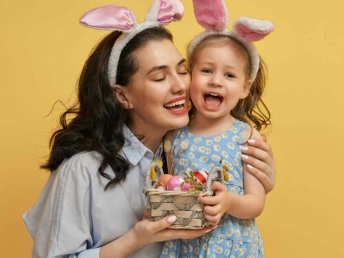 Mãe presenteando sua filha de 1 ano com uma cesta de ovos de cerâmica coloridos por elas mesmas, como alternativa ao chocolate. As duas usam orelhas de coelho brancas; a menina veste um vestido azul com margaridas e a mãe uma camisa social cinza, celebrando a Páscoa com criatividade, carinho e afeto.