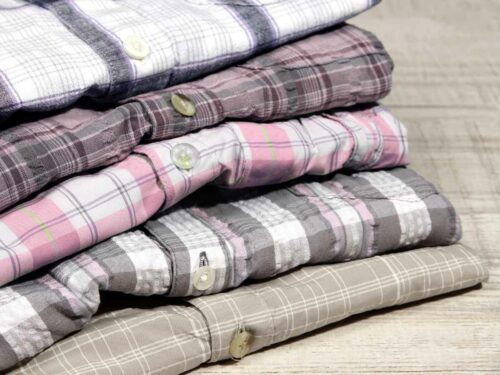 Pilha de camisas xadrez dobradas, organizadas sobre uma superfície de madeira. As peças apresentam diferentes combinações de tons suaves como rosa, cinza, branco e bege.