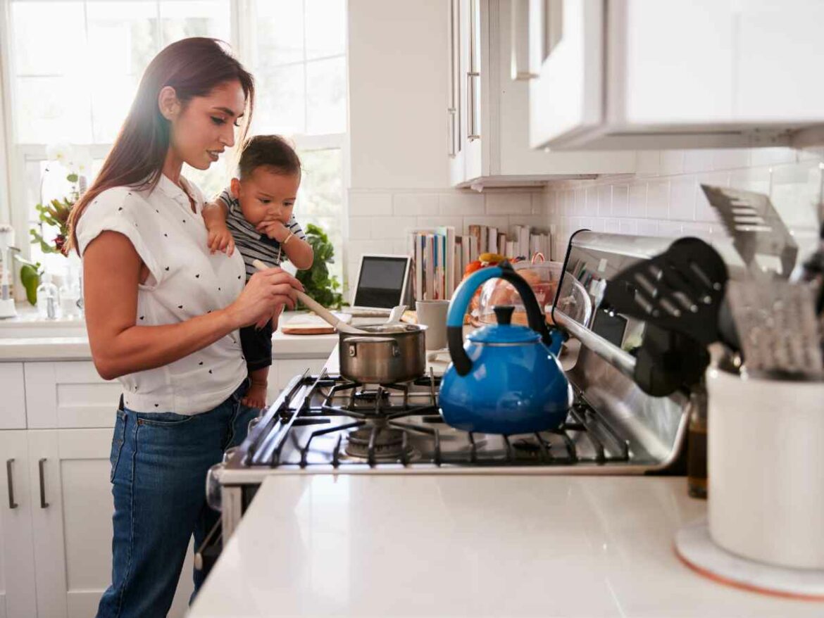 Mãe cozinhando cim seu bebê em seu colo. Na panela que está no fogo, ela faz comidas juninas.