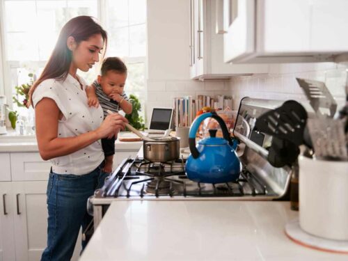Mãe cozinhando com seu bebê em seu colo. Na panela que está no fogo, ela faz comidas juninas.