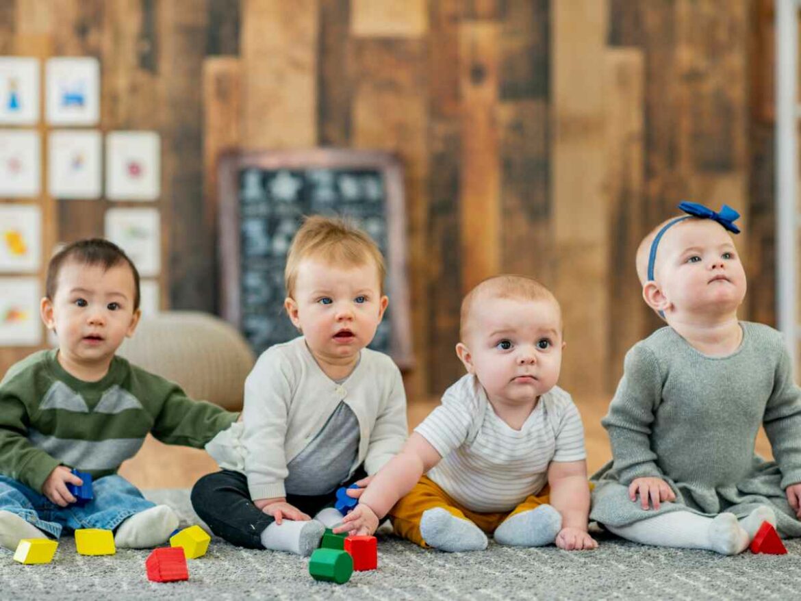 Quatro bebês sentados lado a lado no chão, brincando com blocos coloridos em um ambiente acolhedor. A imagem representa o desenvolvimento da socialização e da empatia nos primeiros anos de vida, por meio da convivência com outras crianças, da observação e da interação, fundamentais para o aprendizado das regras de convivência, respeito e construção de laços saudáveis.