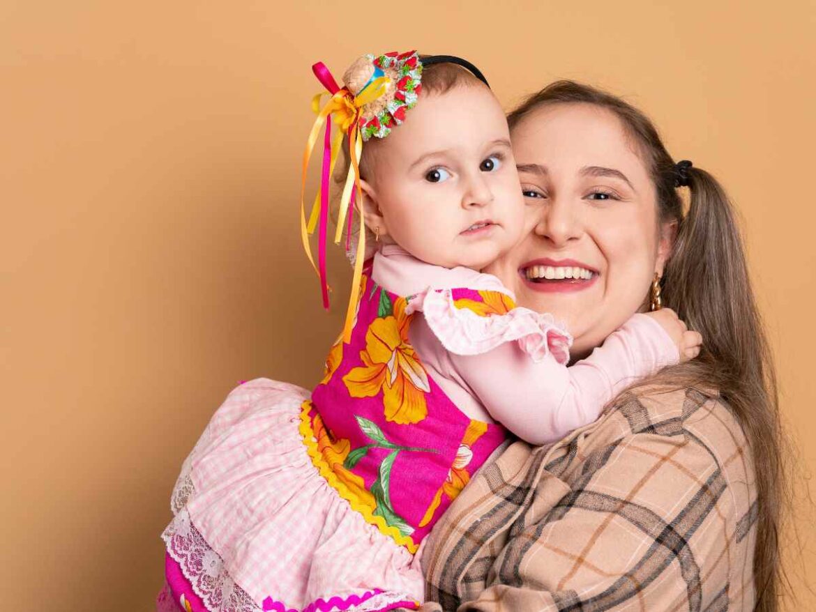 Mãe sorridente segurando sua filha pequena nos braços. A menina está vestida com um look junino, com vestido colorido e adereço na cabeça com fitas, enquanto a mãe usa camisa xadrez.