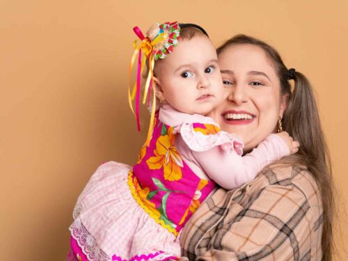 Mãe sorridente segurando sua filha pequena nos braços. A menina está vestida com um look junino, com vestido colorido e adereço na cabeça com fitas, enquanto a mãe usa camisa xadrez.