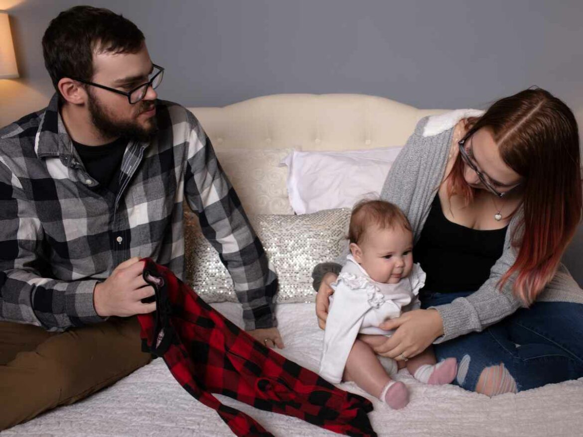 Casal sentado na cama com o bebê, se preparando para trocar sua roupa. O pai segura uma peça de roupa xadrez vermelha e preta, enquanto a mãe segura carinhosamente o bebê, que sorri.