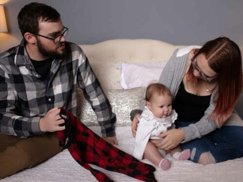 Casal sentado na cama com o bebê, se preparando para trocar sua roupa. O pai segura uma peça de roupa xadrez vermelha e preta, enquanto a mãe segura carinhosamente o bebê, que sorri.