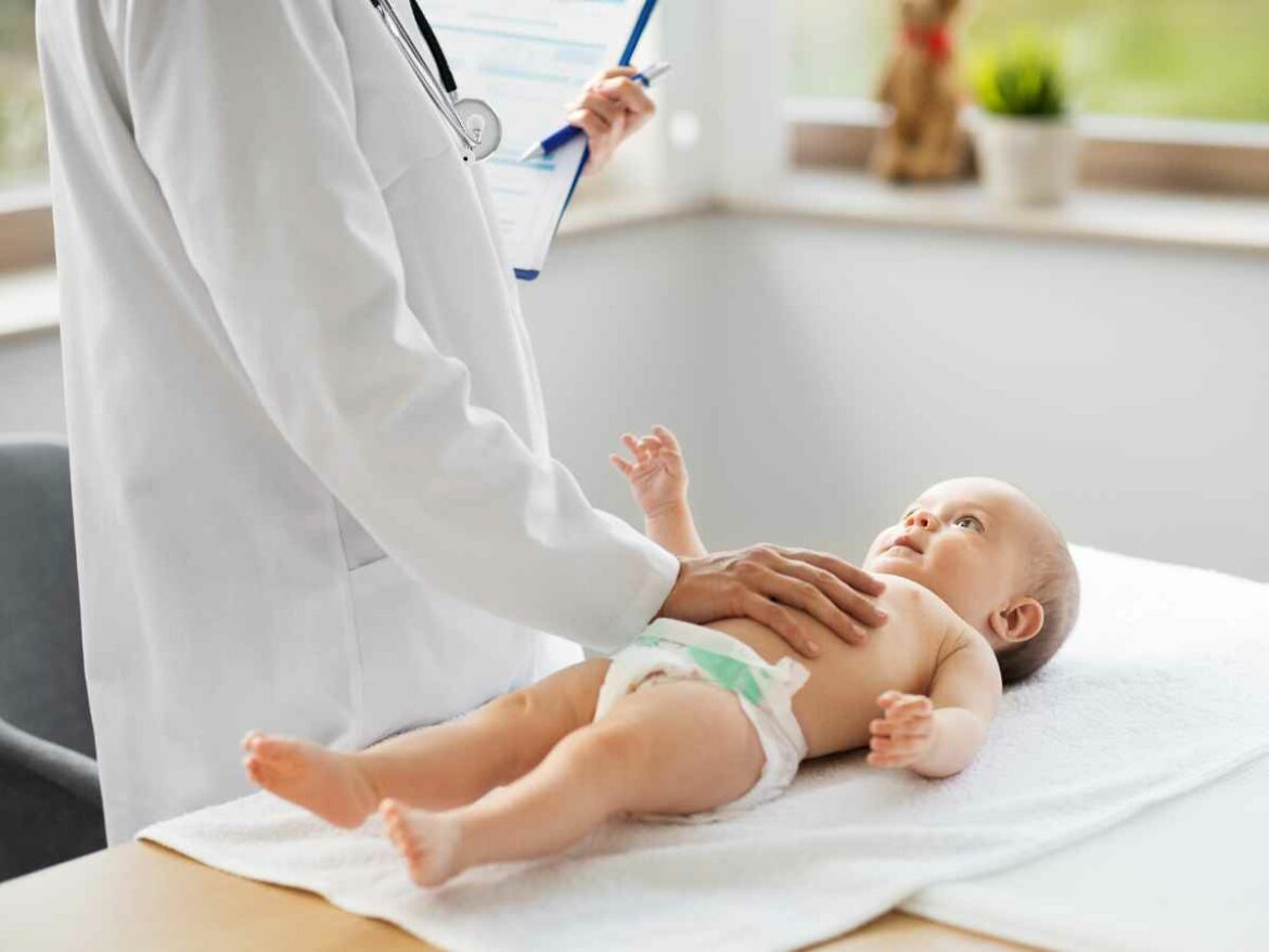 Médico realiza exame físico em bebê deitado em maca com fralda, tocando suavemente sua barriga. A consulta acontece em um ambiente claro e acolhedor.