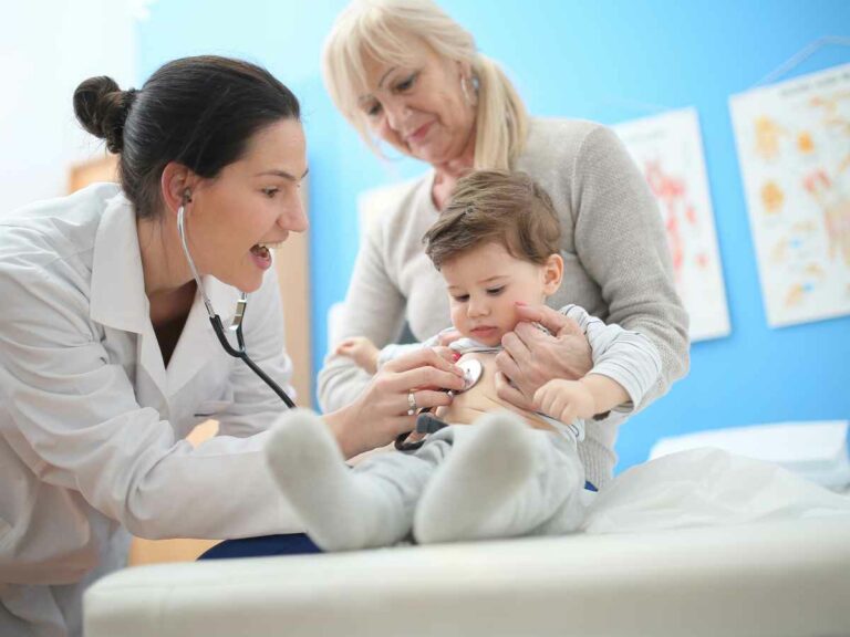 Pediatra examina um bebê com estetoscópio enquanto ele está no colo de uma mulher mais velha, possivelmente a avó.