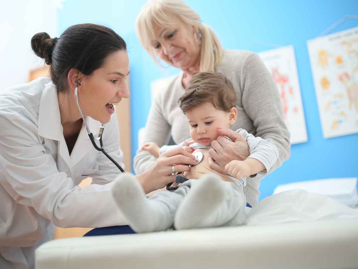 Pediatra examina um bebê com estetoscópio enquanto ele está no colo de uma mulher mais velha, possivelmente a avó.