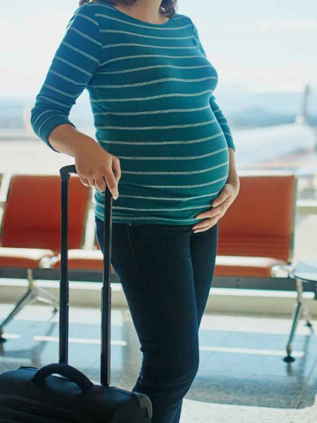 Mulher grávida com uma mão sobre a barriga e outra segurando uma mala de rodinhas, aguardando em um aeroporto com um avião visível ao fundo.