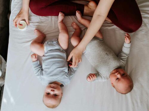 Dois bebês deitados lado a lado em uma cama enquanto um adulto aplica creme em um deles, representando cuidados com a pele dos pequenos em dias frios.