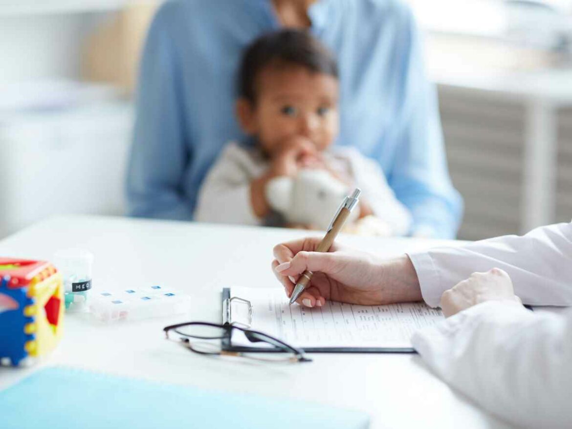 Profissional da saúde preenche formulário em uma consulta, enquanto uma criança pequena senta no colo de um responsável ao fundo.