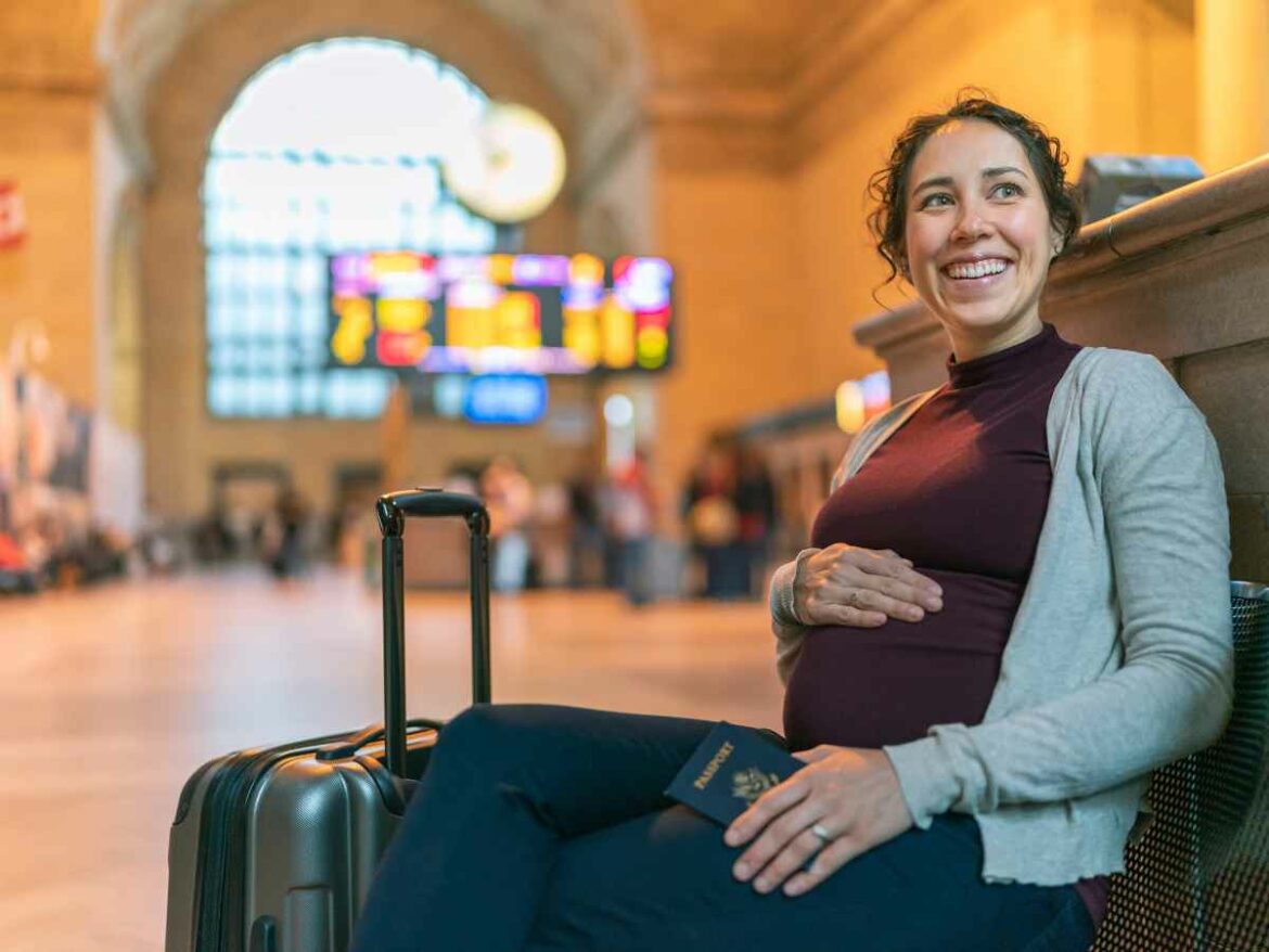 Mulher grávida sentada em terminal de transporte com mala e passaporte na mão, sorrindo enquanto espera.