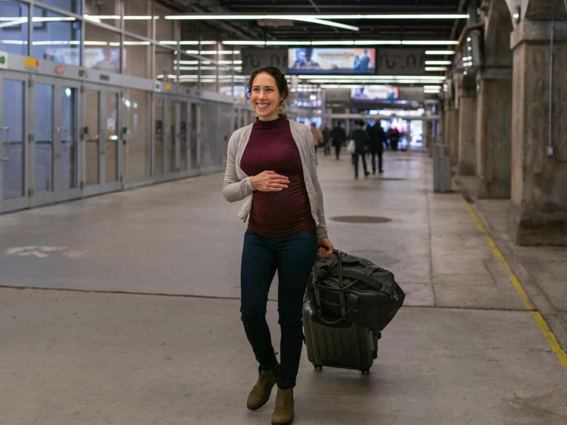 Mulher grávida caminha por uma estação de transporte carregando uma mala de rodinhas.